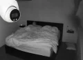 Un'immagine in bianco e nero catturata da una telecamera di sicurezza per la visione notturna mostra due persone distese a letto sotto una coperta in una stanza scarsamente illuminata, probabilmente un Airbnb. Le telecamere sono visibili nell'angolo in alto a sinistra dell'immagine.