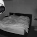 Un'immagine in bianco e nero catturata da una telecamera di sicurezza per la visione notturna mostra due persone distese a letto sotto una coperta in una stanza scarsamente illuminata, probabilmente un Airbnb. Le telecamere sono visibili nell'angolo in alto a sinistra dell'immagine.