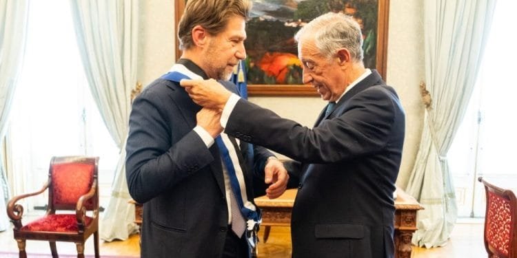 Due uomini in giacca e cravatta sono in piedi in una stanza elegante con mobili decorati e un grande dipinto sullo sfondo. L'Ambasciatore Carlo Formosa è sulla destra, mentre mette con cura una medaglia al collo dell'altro uomo. Entrambi sembrano concentrati sul compito da svolgere.