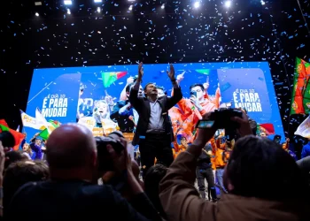 Luis Montenegro sta su un palco con le braccia alzate, dopo aver vinto le Elezioni in Portogallo nel 2024. È circondato da persone. I coriandoli cadono dall'alto. Lo sfondo presenta grandi schermi con immagini e testo, tra cui "E DÍA 10 É PARA MUDAR". La folla tiene bandiere portoghesi e indossa colori vivaci.