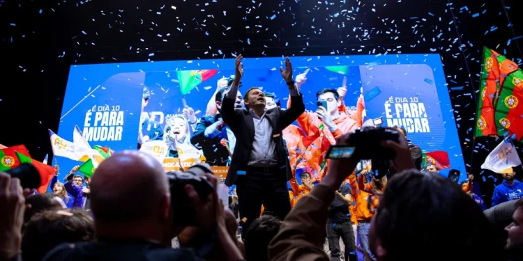Luis Montenegro sta su un palco con le braccia alzate, dopo aver vinto le Elezioni in Portogallo nel 2024. È circondato da persone. I coriandoli cadono dall'alto. Lo sfondo presenta grandi schermi con immagini e testo, tra cui "E DÍA 10 É PARA MUDAR". La folla tiene bandiere portoghesi e indossa colori vivaci.