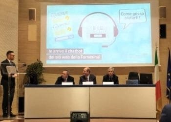 Un uomo sta su un podio e si rivolge a un pubblico attento. Dietro di lui, altri tre uomini siedono a un tavolo sul palco. Su un grande schermo è ben visibile il Chatbot della Farnesina con il testo che recita: "In arrivo il chatbot dei siti web della Farnesina!" e "Come posso aiutarti?".