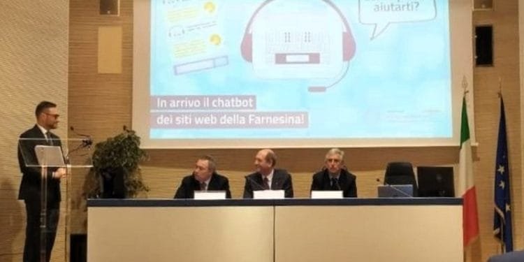 Un uomo sta su un podio e si rivolge a un pubblico attento. Dietro di lui, altri tre uomini siedono a un tavolo sul palco. Su un grande schermo è ben visibile il Chatbot della Farnesina con il testo che recita: "In arrivo il chatbot dei siti web della Farnesina!" e "Come posso aiutarti?".