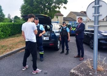Un giovane in abiti casual si trova vicino ad un veicolo della polizia e parla con due agenti in uniforme che chiedono di pagare una multa ricevuta all'estero. Un altro uomo in giacca e cravatta osserva la scena. L'ambientazione sembra essere una zona residenziale con diverse auto della polizia parcheggiate intorno.