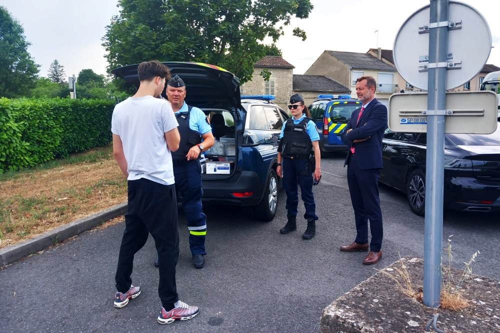 Un giovane in abiti casual si trova vicino ad un veicolo della polizia e parla con due agenti in uniforme che chiedono di pagare una multa ricevuta all'estero. Un altro uomo in giacca e cravatta osserva la scena. L'ambientazione sembra essere una zona residenziale con diverse auto della polizia parcheggiate intorno.