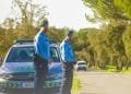 Due agenti della GNR in uniforme stanno sul ciglio di una strada vicino alla loro auto di pattuglia parcheggiata. L'immagine rappresenta i controlli stradali della polizia in portogallo. Sono all'aperto in una giornata soleggiata, con alberi e vegetazione sullo sfondo.