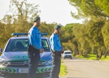 Due agenti della GNR in uniforme stanno sul ciglio di una strada vicino alla loro auto di pattuglia parcheggiata. L'immagine rappresenta i controlli stradali della polizia in portogallo. Sono all'aperto in una giornata soleggiata, con alberi e vegetazione sullo sfondo.