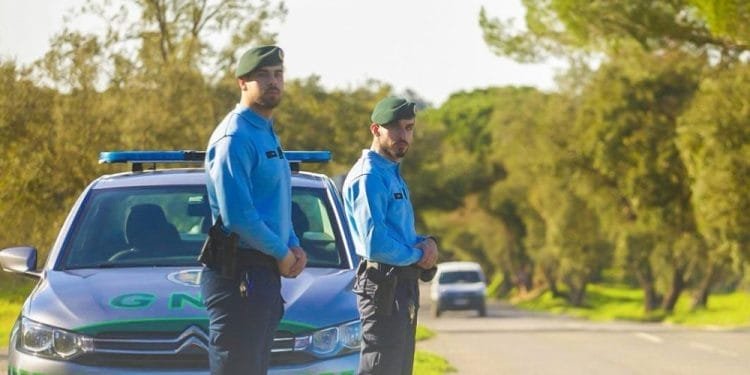 Due agenti della GNR in uniforme stanno sul ciglio di una strada vicino alla loro auto di pattuglia parcheggiata. L'immagine rappresenta i controlli stradali della polizia in portogallo. Sono all'aperto in una giornata soleggiata, con alberi e vegetazione sullo sfondo.
