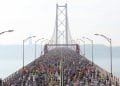 Una grande folla di corridori partecipanti alla mezza maratona Lisbona attraversa un ponte sospeso con un cielo nebbioso sullo sfondo. Il ponte è fiancheggiato da lampioni e all'orizzonte si possono vedere colline lontane e una statua.