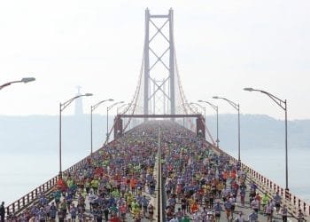 Una grande folla di corridori partecipanti alla mezza maratona Lisbona attraversa un ponte sospeso con un cielo nebbioso sullo sfondo. Il ponte è fiancheggiato da lampioni e all'orizzonte si possono vedere colline lontane e una statua.