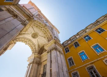 Vista dal basso dell'Arco da Rua Augusta, un grande arco trionfale in pietra a Lisbona, Portogallo, con intricati intagli e statue, contro un cielo azzurro. Adiacente all'arco c'è un edificio giallo brillante con numerose finestre che simboleggiano l'era del regime fiscale privilegiato in architettura.