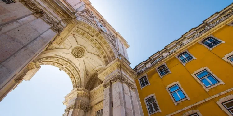 Vista dal basso dell'Arco da Rua Augusta, un grande arco trionfale in pietra a Lisbona, Portogallo, con intricati intagli e statue, contro un cielo azzurro. Adiacente all'arco c'è un edificio giallo brillante con numerose finestre che simboleggiano l'era del regime fiscale privilegiato in architettura.