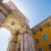 Vista dal basso dell'Arco da Rua Augusta, un grande arco trionfale in pietra a Lisbona, Portogallo, con intricati intagli e statue, contro un cielo azzurro. Adiacente all'arco c'è un edificio giallo brillante con numerose finestre che simboleggiano l'era del regime fiscale privilegiato in architettura.