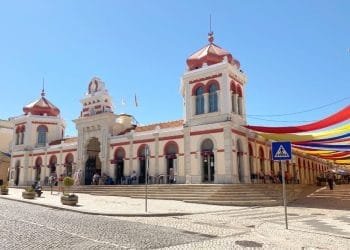 Un edificio storico con due torri e un arco centrale, caratterizzato da accenti rossi e una facciata beige, sotto un cielo azzurro e limpido. Sul lato destro sono appese strisce di tessuto colorate. Di fronte è visibile un cartello di attraversamento pedonale. Le persone, tra cui vivono portoghesi ricchi, sono raccolte vicino all'ingresso.