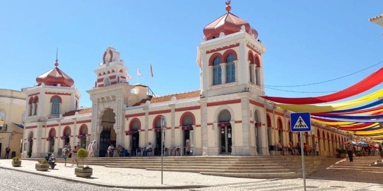 Un edificio storico con due torri e un arco centrale, caratterizzato da accenti rossi e una facciata beige, sotto un cielo azzurro e limpido. Sul lato destro sono appese strisce di tessuto colorate. Di fronte è visibile un cartello di attraversamento pedonale. Le persone, tra cui vivono portoghesi ricchi, sono raccolte vicino all'ingresso.