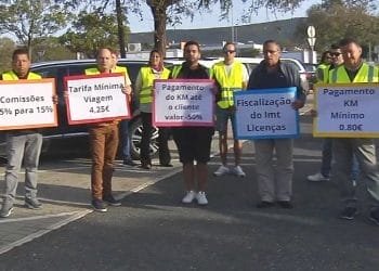 Un gruppo di persone che indossano gilet gialli tengono cartelli di protesta in portoghese. I segnali evidenziano varie richieste relative ai trasporti, come tariffe di commissione, tariffe minime, tariffe per chilometro per i clienti e una maggiore regolamentazione delle licenze che riguardano Autisti TVDE.