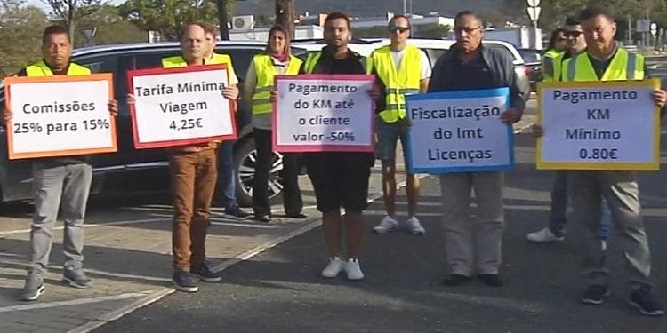 Un gruppo di persone che indossano gilet gialli tengono cartelli di protesta in portoghese. I segnali evidenziano varie richieste relative ai trasporti, come tariffe di commissione, tariffe minime, tariffe per chilometro per i clienti e una maggiore regolamentazione delle licenze che riguardano Autisti TVDE.