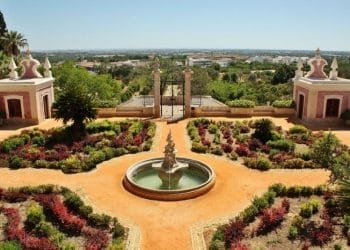 Un giardino formale presenta una fontana centrale circondata da arbusti curati e aiuole geometriche. Al di là del giardino, padiglioni gemelli ornati incorniciano un cancello di ferro, con vista sulla campagna lontana sotto un cielo azzurro e limpido che ricorda il monumento di Estoi villaggio.