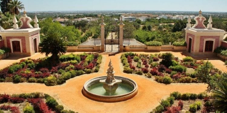 Un giardino formale presenta una fontana centrale circondata da arbusti curati e aiuole geometriche. Al di là del giardino, padiglioni gemelli ornati incorniciano un cancello di ferro, con vista sulla campagna lontana sotto un cielo azzurro e limpido che ricorda il monumento di Estoi villaggio.