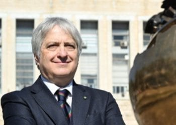 Un individuo dai capelli argentati in abito scuro sta con sicurezza fuori da un edificio con grandi finestre. Posano con le braccia incrociate, indossano una cravatta a righe, con una grande scultura a globo in bronzo parzialmente visibile sulla destra. Claudio Miscia, ambasciatore del Portogallo, emana un'aria di autorità e raffinatezza.