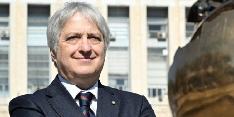 Un individuo dai capelli argentati in abito scuro sta con sicurezza fuori da un edificio con grandi finestre. Posano con le braccia incrociate, indossano una cravatta a righe, con una grande scultura a globo in bronzo parzialmente visibile sulla destra. Claudio Miscia, ambasciatore del Portogallo, emana un'aria di autorità e raffinatezza.