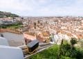 Un tram giallo sale su una ripida collina a Lisbona, Portogallo. La funicolare si muove davanti a edifici bianchi con tetti in terracotta, sullo sfondo di un'ampia vista della città, caratterizzata da un mix di architettura storica e moderna sotto un cielo parzialmente nuvoloso.