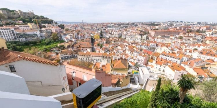 Un tram giallo sale su una ripida collina a Lisbona, Portogallo. La funicolare si muove davanti a edifici bianchi con tetti in terracotta, sullo sfondo di un'ampia vista della città, caratterizzata da un mix di architettura storica e moderna sotto un cielo parzialmente nuvoloso.