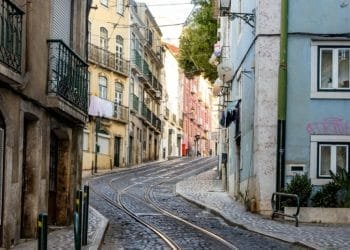 Una strada stretta e acciottolata dove i prezzi delle case a Lisbona sono aumentati, fiancheggiata da edifici a più piani dai colori pastello. I binari del tram corrono in mezzo alla strada. Alcuni edifici sono dotati di balconi con ringhiera e biancheria stesa. La strada sale leggermente e curva in avanti.