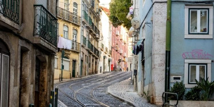 Una strada stretta e acciottolata dove i prezzi delle case a Lisbona sono aumentati, fiancheggiata da edifici a più piani dai colori pastello. I binari del tram corrono in mezzo alla strada. Alcuni edifici sono dotati di balconi con ringhiera e biancheria stesa. La strada sale leggermente e curva in avanti.