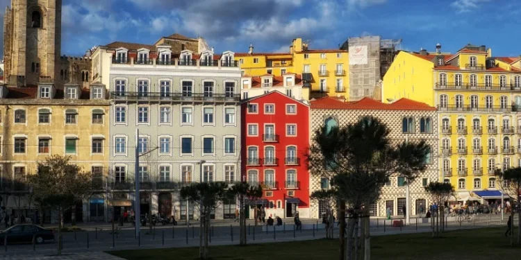 Una fila di colorati edifici a più piani, con facciate rosse, gialle e a motivi geometrici, si trovano in un quartiere luminoso e soleggiato di Lisbona. Sullo sfondo si erge una storica torre in pietra e gli alberi fiancheggiano una piazza aperta pavimentata in primo piano. Il cielo è parzialmente nuvoloso mentre i residenti riflettono sul caso dei prezzi Portogallo.