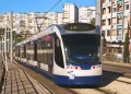 Un moderno tram bianco e blu percorre una strada cittadina, che ricorda il futuro prolungamento della metropolitana di Costa Caparica. Sullo sfondo si trovano edifici residenziali, con diversi appartamenti a molti piani dotati di balconi. Le linee elettriche aeree corrono lungo i binari del tram mentre la luce del giorno illumina la scena.