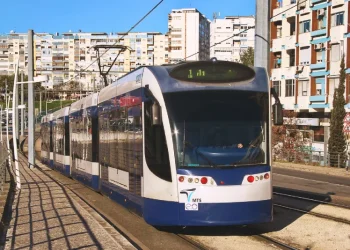 Un moderno tram bianco e blu percorre una strada cittadina, che ricorda il futuro prolungamento della metropolitana di Costa Caparica. Sullo sfondo si trovano edifici residenziali, con diversi appartamenti a molti piani dotati di balconi. Le linee elettriche aeree corrono lungo i binari del tram mentre la luce del giorno illumina la scena.