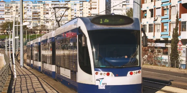Un moderno tram bianco e blu percorre una strada cittadina, che ricorda il futuro prolungamento della metropolitana di Costa Caparica. Sullo sfondo si trovano edifici residenziali, con diversi appartamenti a molti piani dotati di balconi. Le linee elettriche aeree corrono lungo i binari del tram mentre la luce del giorno illumina la scena.