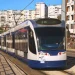 Un moderno tram bianco e blu percorre una strada cittadina, che ricorda il futuro prolungamento della metropolitana di Costa Caparica. Sullo sfondo si trovano edifici residenziali, con diversi appartamenti a molti piani dotati di balconi. Le linee elettriche aeree corrono lungo i binari del tram mentre la luce del giorno illumina la scena.