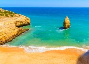 Vista panoramica sulla costa di una spiaggia sabbiosa con acqua turchese e una grande formazione rocciosa che si innalza dal mare vicino alla riva. Scogliere con vegetazione rada incorniciano le spiagge sul lato sinistro sotto un cielo azzurro e limpido, che ricorda la splendida costa dell'Algarve.