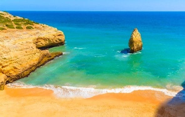 Vista panoramica sulla costa di una spiaggia sabbiosa con acqua turchese e una grande formazione rocciosa che si innalza dal mare vicino alla riva. Scogliere con vegetazione rada incorniciano le spiagge sul lato sinistro sotto un cielo azzurro e limpido, che ricorda la splendida costa dell'Algarve.
