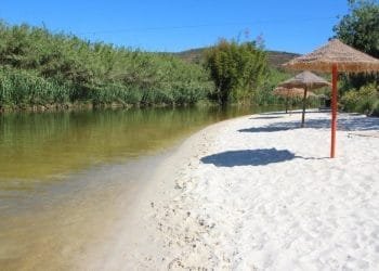 Una spiaggia tranquilla con sabbia bianca e un fiume calmo e poco profondo fiancheggiato da vegetazione. Numerosi ombrelloni di paglia forniscono ombra lungo la battigia. Il cielo è terso e azzurro, suggerendo una giornata soleggiata a 30 gradi. Sullo sfondo sono visibili le colline.