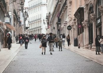 Una vivace scena di strada in una città europea con persone che camminano lungo un'ampia strada acciottolata fiancheggiata da negozi ed edifici storici. La strada, che ricorda quelle dove potresti trovare stanze affitto in Portogallo, è decorata con lampioni decorati e il cielo coperto suggerisce una giornata fresca.