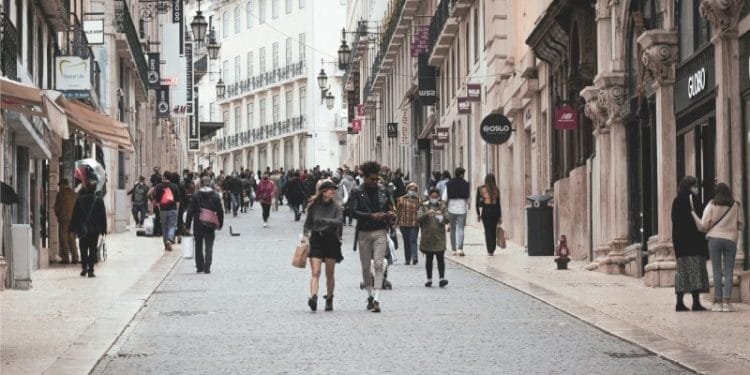 Una vivace scena di strada in una città europea con persone che camminano lungo un'ampia strada acciottolata fiancheggiata da negozi ed edifici storici. La strada, che ricorda quelle dove potresti trovare stanze affitto in Portogallo, è decorata con lampioni decorati e il cielo coperto suggerisce una giornata fresca.