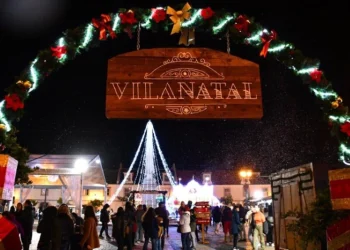 Una scena notturna a Vila Real de Santo Antonio di un festoso mercatino di Natale che mette in mostra un cartello di legno illuminato con la scritta "Vilanatal". Riuniti sotto di esso i visitatori si godono le decorazioni e le luci, con un grande albero di Natale illuminato che fornisce uno sfondo magico.