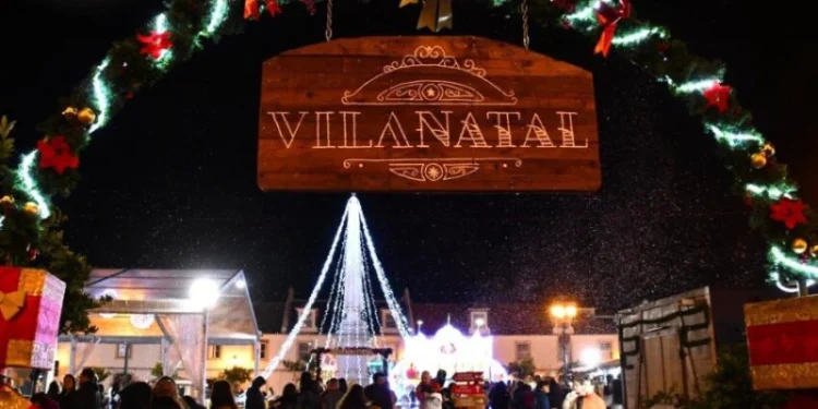 Una scena notturna a Vila Real de Santo Antonio di un festoso mercatino di Natale che mette in mostra un cartello di legno illuminato con la scritta "Vilanatal". Riuniti sotto di esso i visitatori si godono le decorazioni e le luci, con un grande albero di Natale illuminato che fornisce uno sfondo magico.