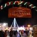 Una scena notturna a Vila Real de Santo Antonio di un festoso mercatino di Natale che mette in mostra un cartello di legno illuminato con la scritta "Vilanatal". Riuniti sotto di esso i visitatori si godono le decorazioni e le luci, con un grande albero di Natale illuminato che fornisce uno sfondo magico.