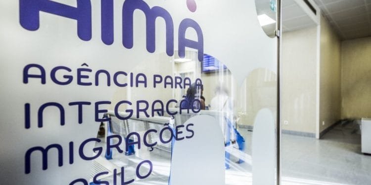 Porta a vetri di un ufficio con la scritta "AIMA Agência para a Integração Migrações e Asilo" in viola. Ti chiedi cosa è AIMA? L'interno, visibile attraverso il vetro, mostra poche persone e un arredamento moderno e minimalista.