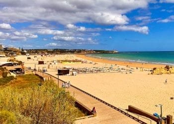 Una vista panoramica di una spiaggia sabbiosa con una passerella in legno che la costeggia. Si possono vedere persone camminare sul lungomare e rilassarsi sulla spiaggia. Il cielo è parzialmente nuvoloso con cieli azzurri e sullo sfondo sono visibili gli edifici lungo l'interessante costa del Portogallo.