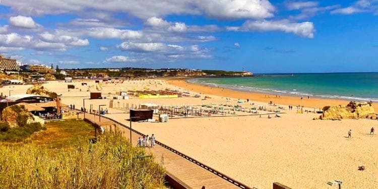 Una vista panoramica di una spiaggia sabbiosa con una passerella in legno che la costeggia. Si possono vedere persone camminare sul lungomare e rilassarsi sulla spiaggia. Il cielo è parzialmente nuvoloso con cieli azzurri e sullo sfondo sono visibili gli edifici lungo l'interessante costa del Portogallo.