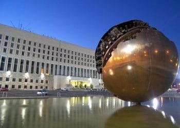 Una grande scultura a sfera dorata, parzialmente decostruita nella parte superiore, si erge in una vasca d'acqua riflettente di fronte al grande edificio della Farnesina a più piani e dotato di numerose finestre. La scena è illuminata da luci artificiali durante il crepuscolo, creando un'atmosfera surreale in stile MIRTA.