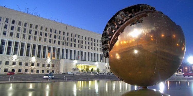 Una grande scultura a sfera dorata, parzialmente decostruita nella parte superiore, si erge in una vasca d'acqua riflettente di fronte al grande edificio della Farnesina a più piani e dotato di numerose finestre. La scena è illuminata da luci artificiali durante il crepuscolo, creando un'atmosfera surreale in stile MIRTA.