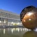 Una grande scultura a sfera dorata, parzialmente decostruita nella parte superiore, si erge in una vasca d'acqua riflettente di fronte al grande edificio della Farnesina a più piani e dotato di numerose finestre. La scena è illuminata da luci artificiali durante il crepuscolo, creando un'atmosfera surreale in stile MIRTA.