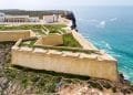 Veduta aerea della storica Fortezza di Sagres in Portogallo, uno dei grandi monumenti dell'algarve, adagiato su un promontorio roccioso circondato dall'Oceano Atlantico. La fortezza presenta mura giallastre, vegetazione verde ed edifici all'interno, con onde che si infrangono contro le scogliere sottostanti.
