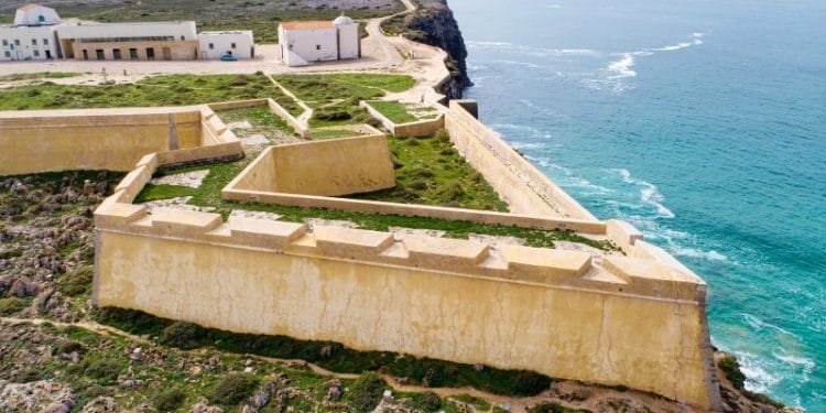 Veduta aerea della storica Fortezza di Sagres in Portogallo, uno dei grandi monumenti dell'algarve, adagiato su un promontorio roccioso circondato dall'Oceano Atlantico. La fortezza presenta mura giallastre, vegetazione verde ed edifici all'interno, con onde che si infrangono contro le scogliere sottostanti.